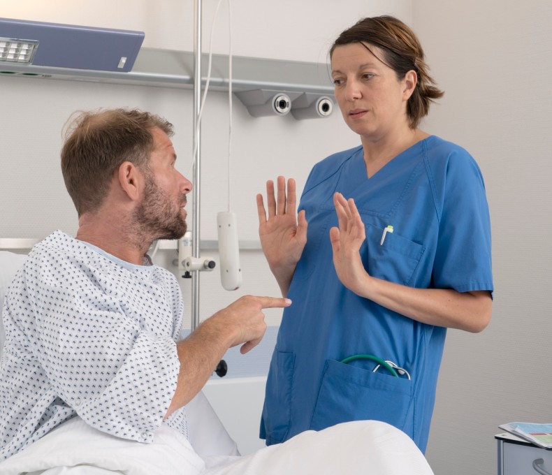 Eine Pflegerin weicht vor einem Patienten zurück, der mit dem Finger auf sie zeigt. Sie hebt abwehrend die Hände. Die Pflegerin trägt einen blauen Kittel. Der Patient ein Krankenhausnachthemd. Eine Pflegerin weicht vor einem Patienten zurück, der mit dem Finger auf sie zeigt. Sie hebt abwehrend die Hände. Die Pflegerin trägt einen blauen Kittel. Der Patient ein Krankenhausnachthemd.